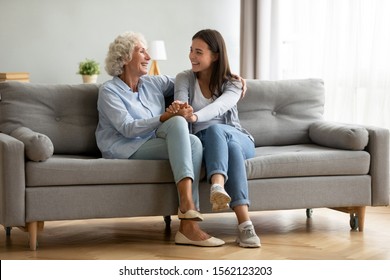 Happy adult granddaughter and senior grandmother having fun enjoying talk sit on sofa in modern living room, smiling old mother hugging young grown daughter bonding chatting relaxing at home together