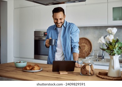 A happy adult man grins and reads news on a tablet with a cup of coffee in his hand in a kitchen. - Powered by Shutterstock