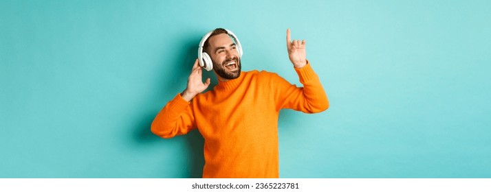 Happy adult man in orange sweater, looking up and listening music in headphones, standing over blue background. - Shutterstock ID 2365223781