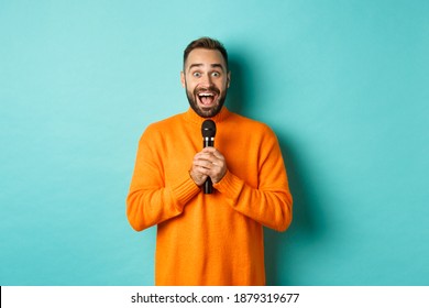 Happy adult man singing karaoke, holding microphone and looking at camera, standing in orange sweater against turquoise background