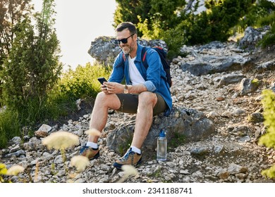 Happy adult man sitting on rock using his phone while hiking