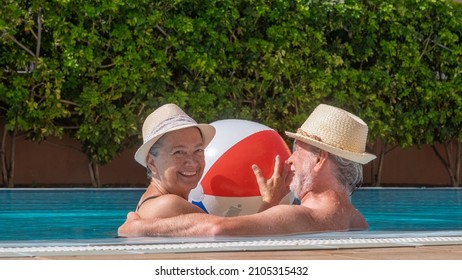 Happy adult mature senior caucasian couple laughing looking at camera floating in outdoor swimming pool playing with inflatable balloon. Active elderly people enjoying healthy lifestyle and retirement