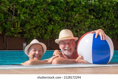 Happy adult mature senior caucasian couple laughing looking at camera floating in outdoor swimming pool playing with inflatable balloon. Active elderly people enjoying healthy lifestyle and retirement