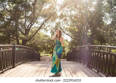 Happy adult middle-aged woman enjoying summer on park walks across bridge outdoors, dressed in green fashionable dress, looking away. Outdoor portrait of middle aged blonde female model. Copy space