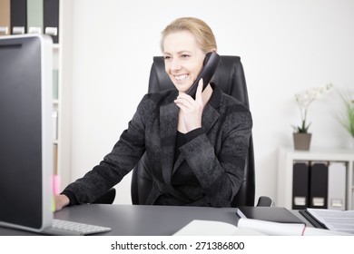 Happy Adult Office Woman At Her Office Talking To Someone On Telephone While Looking At Her Computer Screen.