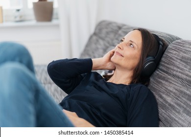 Happy Adult Woman Listening To Black Headphones While Leaning Back On Dark Grey Couch And Wearing Black Shirt