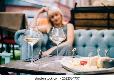 Happy adult woman sitting on couch in bar outdoors with wine glasses and blurry restaurant background scene, drinking white wine and eating cheese. Summer sunny day on patio. People lifestyle