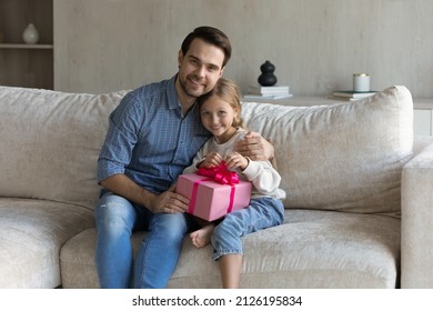 Happy Affectionate Dad Giving Surprise Birthday Gift To Sweet Little Daughter, Hugging Kid On Sofa With Love, Tenderness, Looking At Camera, Smiling, Girl Holding Pink Present Wrap. Family Portrait