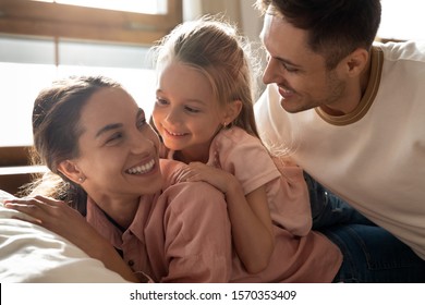 Happy affectionate family young adult parents and cute little girl daughter relax bonding lying on comfortable bed, mum dad laughing playing with small child cuddling together in bedroom in morning