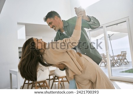 Happy affectionate loving middle aged mature couple dancing at home in kitchen. Smiling older senior man and woman in love having fun at home enjoying dance lit with sunlight. Authentic photo.