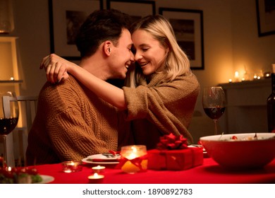 Happy Affectionate Young Couple In Love Hugging Laughing, Drinking Wine, Celebrating Valentines Day Dining Together, Having Romantic Dinner Date In Candle Light Sit At Home Table On Valentine's Day.