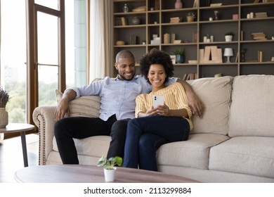 Happy African 30s Couple Sit On Sofa With Cell Phone Smiling Look At Cell Phone Screen Making Video Call With Friends, Using New Mobile Application, Watch Videos, Take Selfie Pictures Relaxing At Home