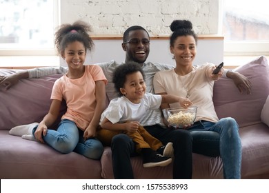 Happy African America Parents Sit On Comfortable Couch In Living Room With Little Kids Watch TV Eat Popcorn Together, Smiling Biracial Family With Children Enjoy Home Weekend Enjoying Movie