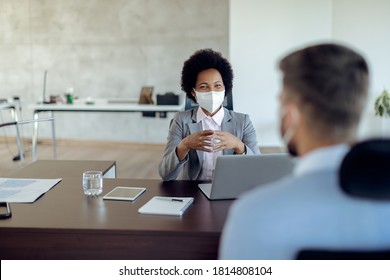 Happy African American Bank Manager Talking To Her Client And Wearing A Face Mask In The Office Due To Coronavirus Epidemic. 