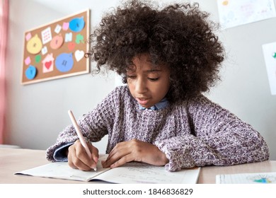 Happy african american black preteen school girl studying in classroom sitting at desk. Smart cute mixed race kid primary school student writing in exercise book doing homework, learning at home.