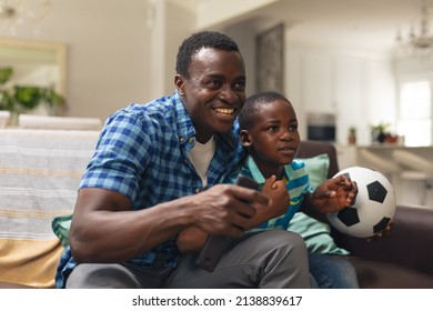 Happy African American Boy And Father With Soccer Ball Sitting On Sofa At Home. Unaltered, Lifestyle, Sport, Fans, Family, Watching Tv, Soccer Match, Cheering And Leisure Concept.