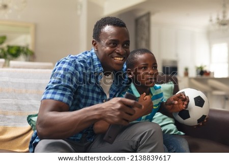 Happy african american boy and father with soccer ball sitting on sofa at home. unaltered, lifestyle, sport, fans, family, watching tv, soccer match, cheering and leisure concept.