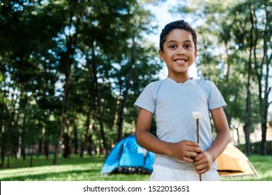 happy african american boy holding stick with sweet marshmallow near camps - Powered by Shutterstock