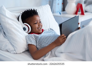 Happy african american boy patient in headphones using tablet in hospital bed. Hospital, medical and healthcare services. - Powered by Shutterstock