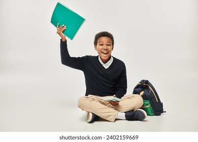 happy african american boy in uniform holding smartphone and notebook while sitting near backpack - Powered by Shutterstock