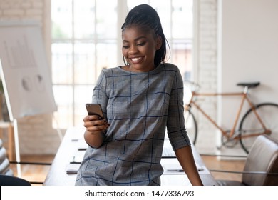 Happy african american business woman holding smartphone in modern office, smiling black female professional texting sms using apps enjoy work with mobile technology online stand in corporate space