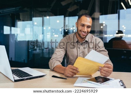Happy african american businessman financier reading letter from bank, man received envelope with notification, man smiling and happy working inside modern office building with laptop on paper work.