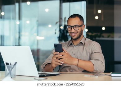 Happy african american businessman in glasses working inside modern office with laptop, man in shirt sitting at table browsing online pages using smartphone. - Powered by Shutterstock