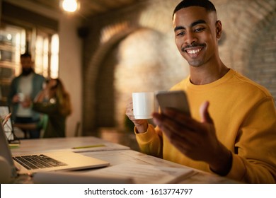 Happy African American businessman reading text message on mobile phone and drinking coffee while working late in the office.  - Powered by Shutterstock