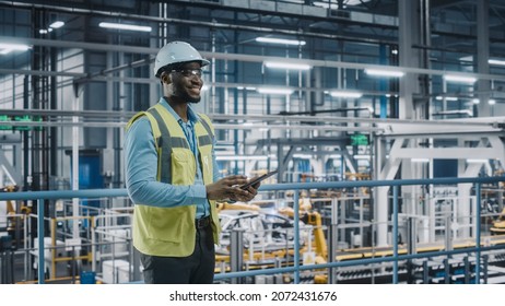 Happy African American Car Factory Engineer In High Visibility Vest Using Tablet Computer. Automotive Industrial Facility Working On Vehicle Production On Automated Technology Assembly Plant.