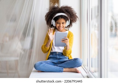 Happy african american child cute preteen girl with bushy hair sitting on windowsill at home, using wireless headphones and modern digital tablet, having online party with friends, copy space - Powered by Shutterstock