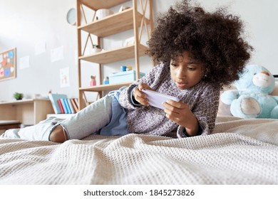 Happy African American Child Kid Girl Holding Phone Lying On Bed In Her Room. Cute Mixed Race Child Using Social Media Mobile Apps On Smartphone, Watching Videos On Cellphone Relaxing In Bedroom.