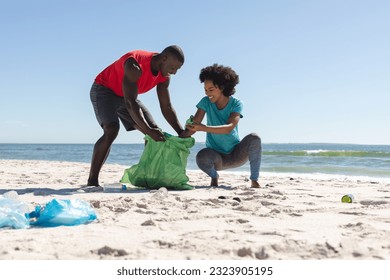 Happy african american couple cleaning sunny beach and collecting litter. Summer, lifestyle, free time, ecology, recycling and vacation, unaltered. - Powered by Shutterstock