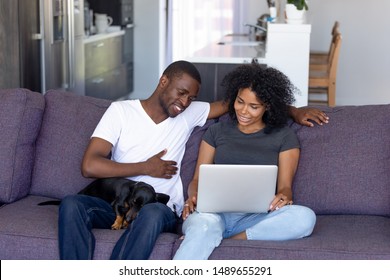 Happy African American Couple With Dackel Dog Relax Sitting On Cozy Sofa At Home Watching Movie On Laptop, Black Man And Woman Rest On Couch With Cute Pet, Browsing Using Computer, Shopping Online