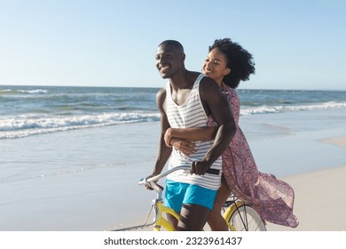 Happy african american couple embracing riding bike on sunny beach by sea. Summer, romance, freedom, relaxation, togetherness and vacation, unaltered. - Powered by Shutterstock