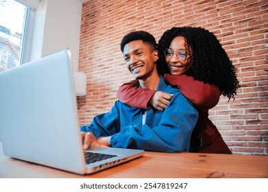 Happy African American couple enjoying time together at home, using a laptop on a cozy desk, embracing and smiling in a warm, relaxed environment. Young people booking their vacations on internet - Powered by Shutterstock