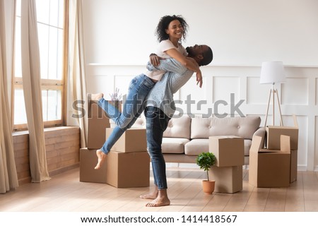 Happy african american couple first time home buyers celebrate new house purchase on moving day, smiling husband embracing lifting excited young wife laughing standing among boxes in own flat house