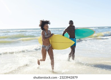 Happy african american couple holding surfboards running out of sea on sunny beach. Summer, togetherness, healthy lifestyle, sport, hobbies, surfing and vacation, unaltered. - Powered by Shutterstock