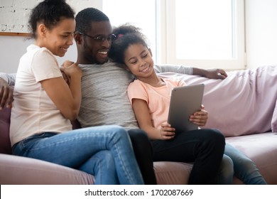 Happy African American Couple Looking At Tablet Screen With Daughter. Diverse Smiling Family Of Mother, Father And Girl Sitting On Couch Using Mobile Device And Gadget Together.