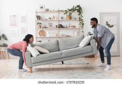 Happy African American Couple Moving Sofa In Living Room, Replacing Furniture At Home, Side View