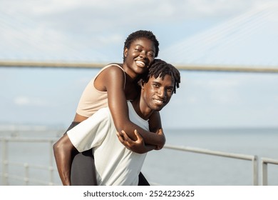 A happy African American couple shares a playful moment by the water, with the sun shining brightly. The woman smiles broadly while being playfully carried on the man back, surrounded by scenic view. - Powered by Shutterstock