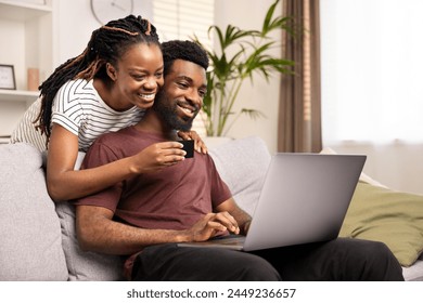 Happy African American Couple Shopping Online Together At Home On Laptop. Love, Technology, and Leisure Concept. Smiling Woman Holding Credit Card Behind Cheerful Man On Computer. - Powered by Shutterstock