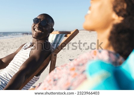 Happy african american couple in sunglasses sitting in deckchairs laughing on sunny beach. Summer, relaxation and vacation, unaltered.