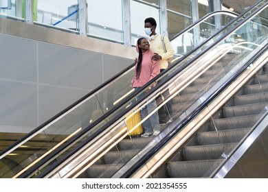 Happy African American Couple Travel In Covid-19 Lockdown. Black Man Wearing Medical Mask Hug Woman Holding Suitcase On Escalator Arriving From Flight. Tourism And Transport In Coronavirus Quarantine