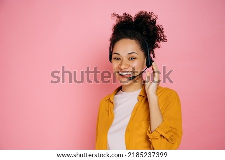 Happy african american curly haired young woman with headset, help desk worker, call center operator, consultant, talking with client, stand on isolated pink background, looks at camera,smile