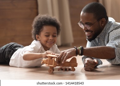 Happy African American Dad And Cute Little Child Boy Play With Wooden Toy Plane Lay On Floor, Funny Small Kid Son And Black Father Having Fun Fly On Airplane At Home, Children Daycare Lifestyle Game