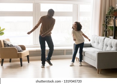 Happy African American Dad And Kid Daughter Laughing Dancing In Living Room Together, Cheerful Cute Child Girl Having Fun With Father Moving To Music Playing At Home, Black Family Funny Activity