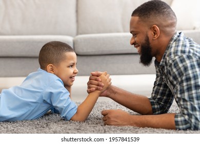 Happy African American Dad And Kid Son Arm Wrestling Competing Lying On Floor At Home. Happy Young Daddy And Little Boy Armwrestling Having Fun Together. Side View