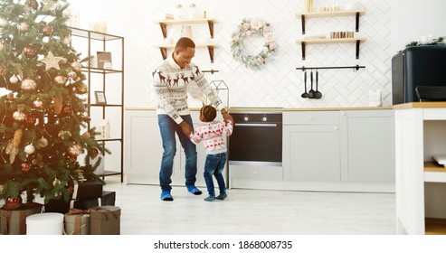 Happy African American Dad And Little Kid Listen Music And Dancing In Modern Cozy House Kitchen Decorated With Xmas Tree Twinkle Lights On Christmas Eve. Happy Family Enjoy Free Time, Holidays Concept