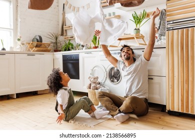 Happy african american dad and son smiling and tossing towels in air while sitting on floor near washing machine during household routine in kitchen at home - Shutterstock ID 2138566895