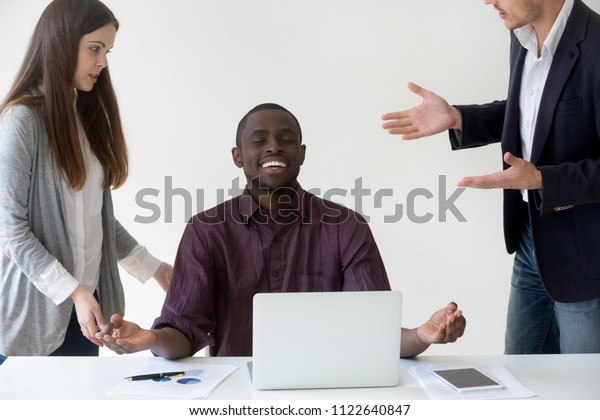 Happy African American employee meditating at\
desk, reaching nirvana state ignoring colleagues talking to him,\
black worker abstracting from annoying colleagues meditating.\
Stress relief concept